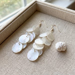 Könnte beinhalten: Hängeohrringe mit mehreren, schimmernden, runden, weißen Muschelstücken. Die Ohrringe haben goldfarbene Haken. Rechts von den Ohrringen liegt eine kleine, weiße Muschel auf einer beigen Oberfläche.