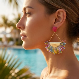 May include: A statement earring featuring a small, round, textured pink stud connected to a gold-tone triangular frame. A semi-circular gold-tone metal piece sits at the base of the triangle, adorned with a vibrant fringe of multi-coloured thread tassels.