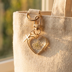 May include: A gold-toned metal keychain featuring a heart-shaped pendant with a clear resin center containing a pressed white flower. A small gold-toned cross charm is attached to the jump ring. The keychain is clipped to a beige canvas tote bag.