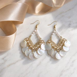 Pode incluir: Um par de brincos dourados com um design de lustre. Cada brinco apresenta uma filigrana em forma de diamante, adornada com pequenas pérolas e detalhes de concha brancos circulares pendurados. Os brincos pendem de ganchos dourados.