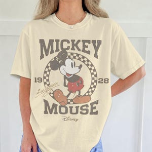 May include: Cream-colored t-shirt featuring a distressed graphic of Mickey Mouse. The design includes the words "Mickey Mouse" in a vintage font, the years "1928" and the Disney logo. The shirt has short sleeves and a relaxed fit.