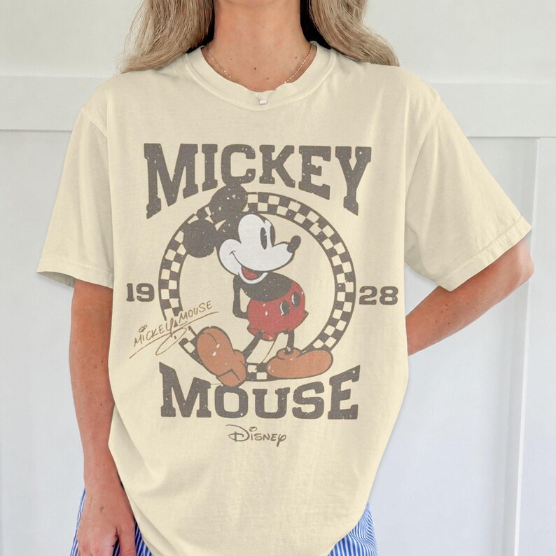 May include: Cream-colored t-shirt featuring a distressed graphic of Mickey Mouse. The design includes the words "Mickey Mouse" in a vintage font, the years "1928" and the Disney logo. The shirt has short sleeves and a relaxed fit.