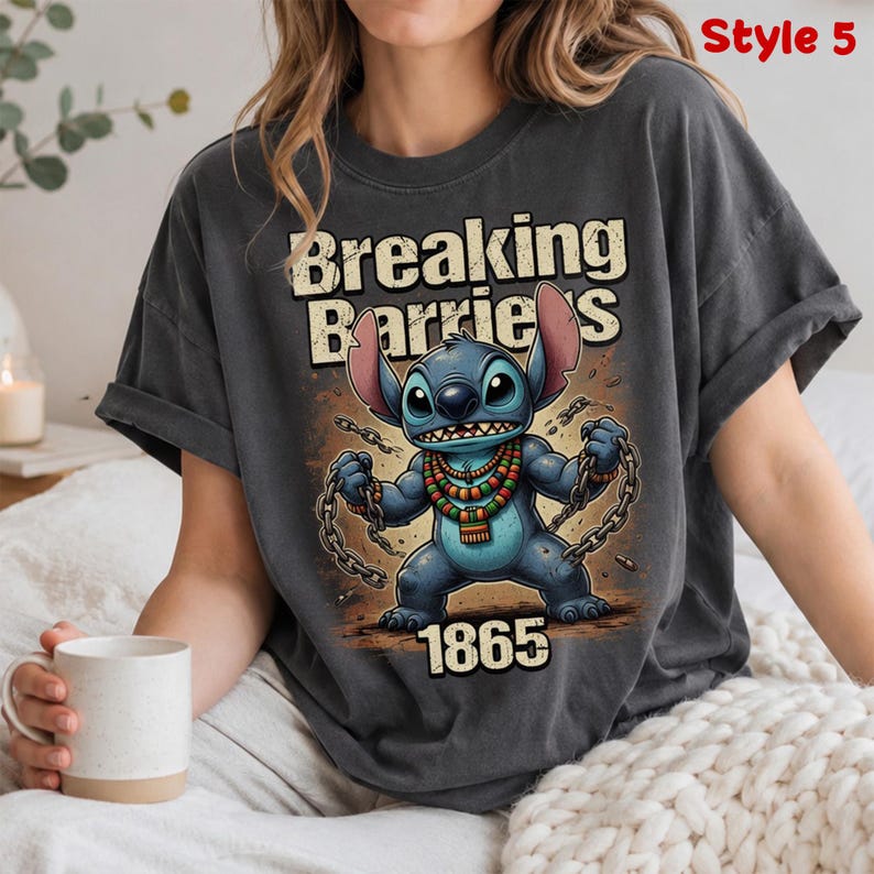 May include: Dark gray t-shirt featuring a cartoon character with the text "Breaking Barriers" and the number "1865". The character is blue with large ears and is holding chains. The shirt has rolled-up sleeves.