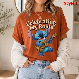 May include: Rust-colored t-shirt featuring a cartoon character playing a ukulele. The character is blue with large ears and a wide smile. The text "Celebrating My Roots" is arched above the character. The shirt is labeled "Style 1".