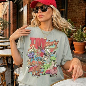 May include: A sage green t-shirt featuring a graphic print of Toy Story characters including Jessie, Woody, Buzz Lightyear, Slinky Dog, Bullseye, Rex, and Hamm. The text reads Toy Story, Jessie Since 1995, Woody, Buzz, Slinky Dog, Bullseye, and Hamm.