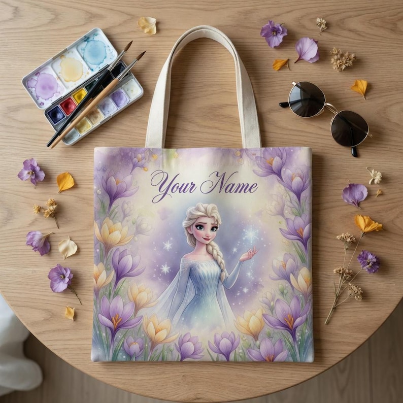 Op de afbeelding: Een canvas draagtas met een illustratie van een prinses in een blauwe jurk, omringd door paarse en gele bloemen. De tekst "Your Name" staat bovenaan. De tas heeft cr&egrave;mekleurige handvatten. Aquarelverf en penselen liggen op de tafel.