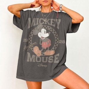 May include: Dark gray t-shirt featuring a distressed graphic of Mickey Mouse. The design includes the words "Mickey Mouse," "1928," and "Disney." Mickey is depicted in his classic pose, wearing red shorts and large yellow shoes. The shirt has a vintage, worn look.