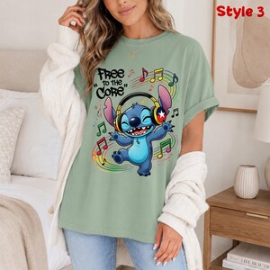 May include: Sage green t-shirt featuring a cartoon character wearing headphones and dancing amidst musical notes. The text "Free to the Core" is above the character. The shirt is a casual style.