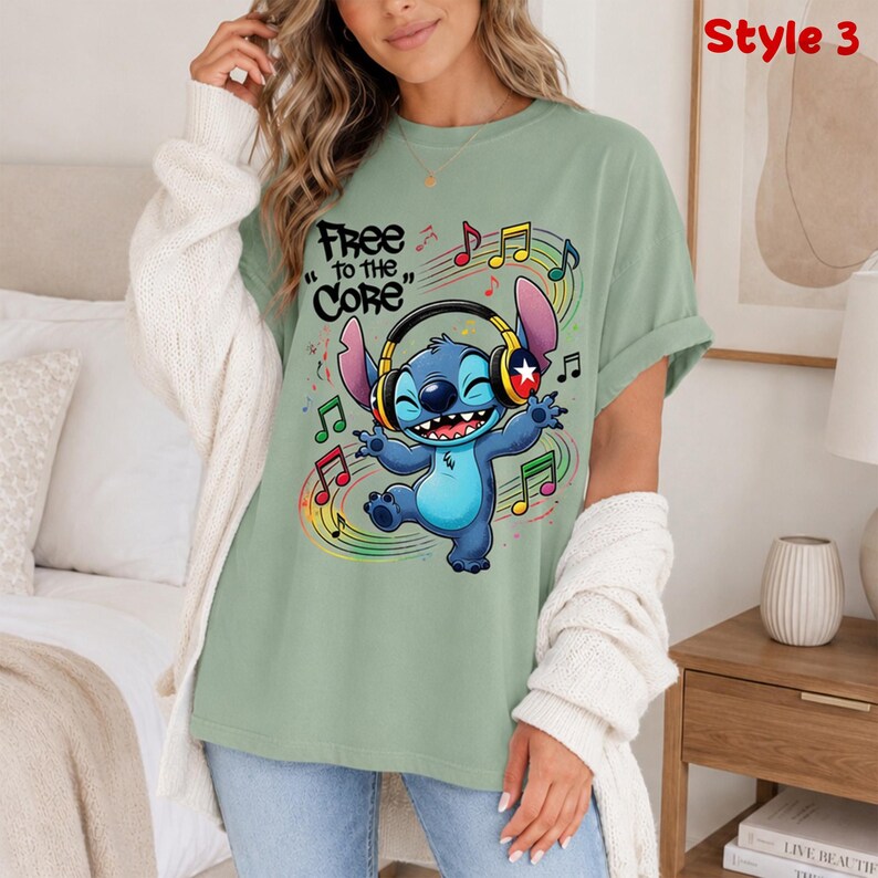 May include: Sage green t-shirt featuring a cartoon character wearing headphones and dancing amidst musical notes. The text "Free to the Core" is above the character. The shirt is a casual style.