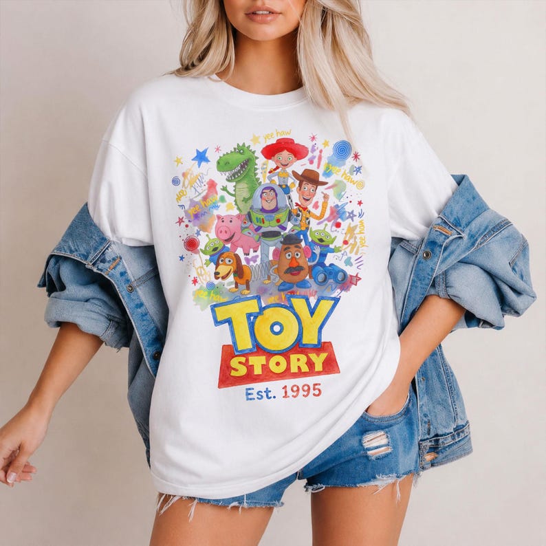 以下が含まれることがあります： カラフルなトイ・ストーリーのキャラクターが描かれた白いTシャツ。ウッディ、バズ・ライトイヤー、その他のキャラクターがデザインされており、「Toy Story」と「Est. 1995」の文字が太字で表示されています。カジュアルで快適なスタイルです。