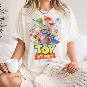 以下が含まれることがあります： クリーム色のTシャツ。トイ・ストーリーのキャラクター、ウッディ、バズ・ライトイヤー、レックスなどのカラフルなグラフィックが特徴です。「TOY STORY」の文字が大きく太字で表示され、その下に「Est. 1995」が赤い長方形で表示されています。ゆったりとしたフィット感のシャツです。