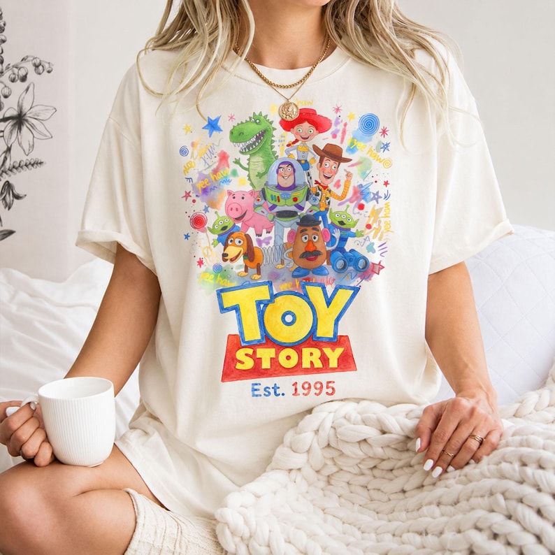 以下が含まれることがあります： クリーム色のTシャツ。トイ・ストーリーのキャラクター、ウッディ、バズ・ライトイヤー、レックスなどのカラフルなグラフィックが特徴です。「TOY STORY」の文字が大きく太字で表示され、その下に「Est. 1995」が赤い長方形で表示されています。ゆったりとしたフィット感のシャツです。