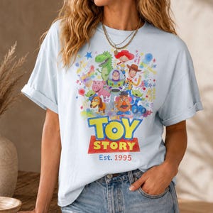 以下が含まれることがあります： 明るい青色のTシャツ。映画「トイ・ストーリー」のキャラクターがカラフルに描かれています。ウッディ、バズ・ライトイヤー、その他のキャラクターがデザインされ、「TOY STORY」と「Est. 1995」の文字が太字で表示されています。