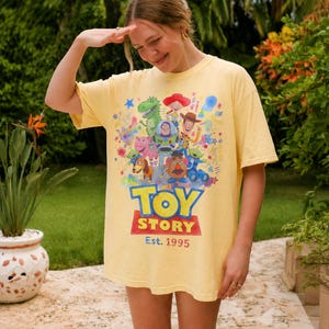 以下が含まれることがあります： 明るい黄色のTシャツ。カラフルなトイ・ストーリーのキャラクターが描かれています。「TOY STORY」の文字は青と赤で、「Est. 1995」が下にあります。ゆったりとしたフィット感のTシャツです。