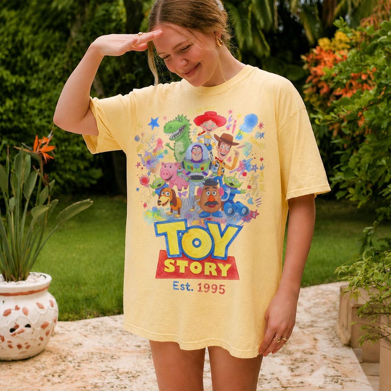 以下が含まれることがあります： 明るい黄色のTシャツ。カラフルなトイ・ストーリーのキャラクターが描かれています。「TOY STORY」の文字は青と赤で、「Est. 1995」が下にあります。ゆったりとしたフィット感のTシャツです。