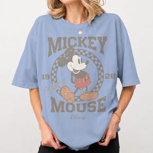 May include: Light blue t-shirt featuring a distressed graphic of Mickey Mouse. The design includes the words "Mickey Mouse" and the year "1928" in a vintage style. The Disney logo is at the bottom of the graphic.