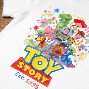 以下が含まれることがあります： 白いTシャツに、バズ・ライトイヤーやウッディなど、トイ・ストーリーのキャラクターが描かれたカラフルなグラフィック。トイ・ストーリーのロゴは黄色と赤で表示され、下に「Est. 1995」と書かれています。デザインには「yee haw」という言葉が含まれています。