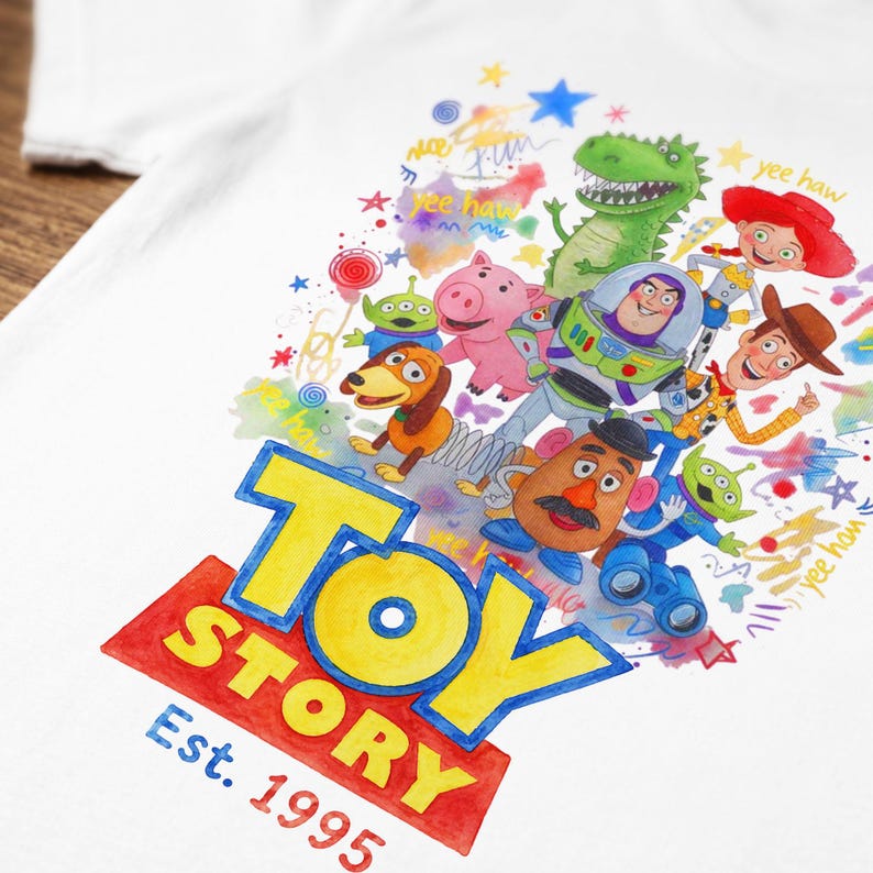 以下が含まれることがあります： 白いTシャツに、バズ・ライトイヤーやウッディなど、トイ・ストーリーのキャラクターが描かれたカラフルなグラフィック。トイ・ストーリーのロゴは黄色と赤で表示され、下に「Est. 1995」と書かれています。デザインには「yee haw」という言葉が含まれています。