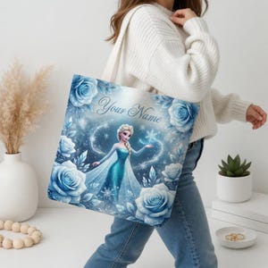 Op de afbeelding: Een blauwe draagtas met een prinsessenillustratie, blauwe rozen en sneeuwvlokken. De tekst "Your Name" staat erop. De tas heeft een witte band. De persoon draagt een witte trui en een blauwe jeans.