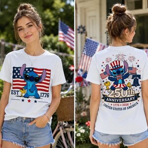 Op de afbeelding: Een wit t-shirt met een blauw alien-personage op de voorkant, een Amerikaanse vlag en de tekst EST 1776. De achterkant toont hetzelfde personage met een patriottische hoed, vuurwerk, de Liberty Bell en 250th ANNIVERSARY 1776-2026 tekst.