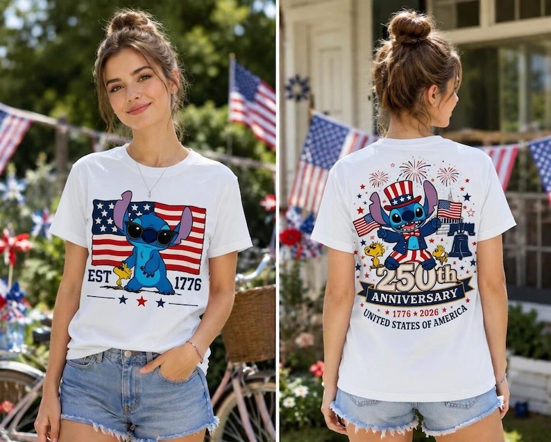 Op de afbeelding: Een wit t-shirt met een blauw alien-personage op de voorkant, een Amerikaanse vlag en de tekst EST 1776. De achterkant toont hetzelfde personage met een patriottische hoed, vuurwerk, de Liberty Bell en 250th ANNIVERSARY 1776-2026 tekst.