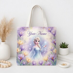 Op de afbeelding: Een witte draagtas met een aquarel illustratie van een prinses in een blauwe jurk, omringd door paarse en gele bloemen. De tekst "Your Name" staat boven de prinses. De tas heeft witte handvatten.