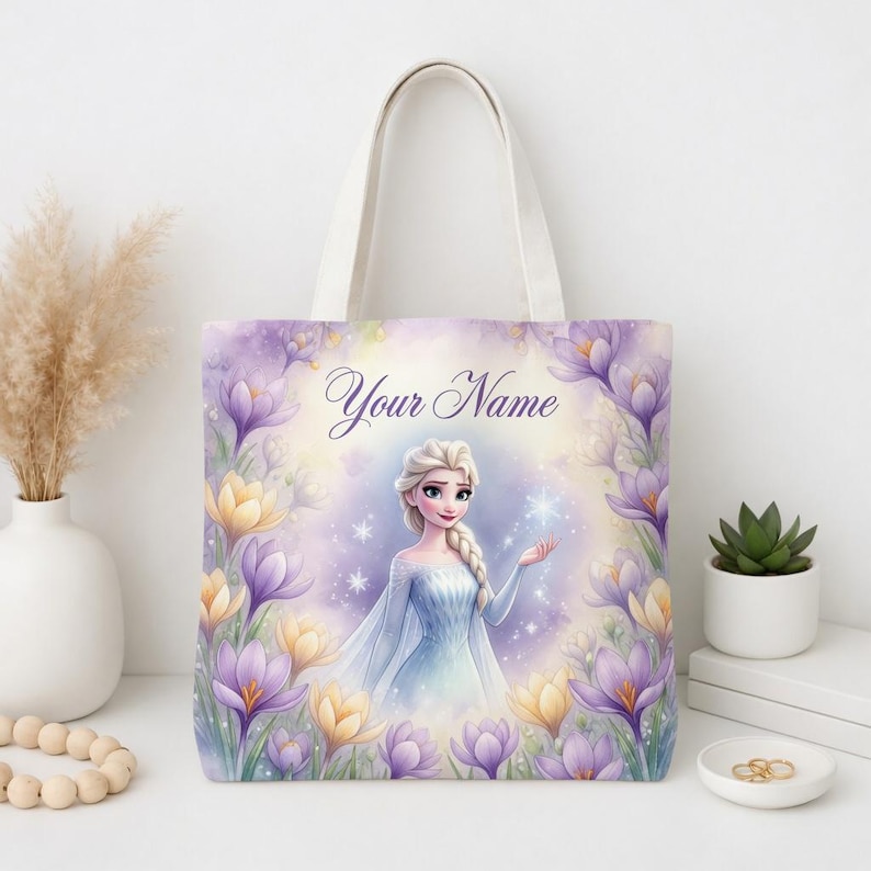 Op de afbeelding: Een witte draagtas met een aquarel illustratie van een prinses in een blauwe jurk, omringd door paarse en gele bloemen. De tekst "Your Name" staat boven de prinses. De tas heeft witte handvatten.