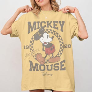 May include: A yellow t-shirt featuring Mickey Mouse. The design includes the words "Mickey Mouse" in a distressed font, along with the years "19" and "28". Mickey Mouse is depicted in a classic pose within a checkered circle. The Disney logo is at the bottom.