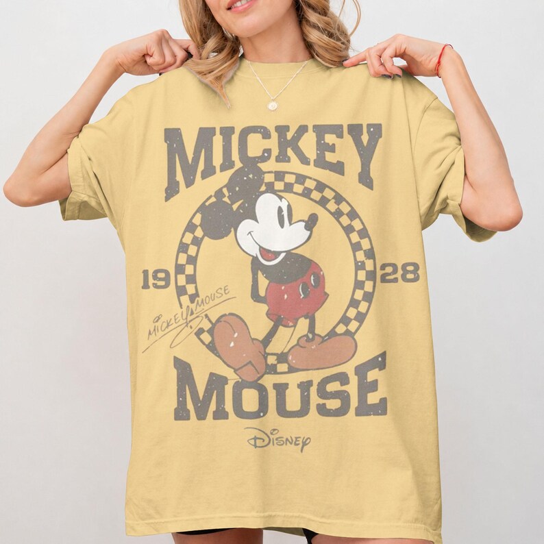 May include: A yellow t-shirt featuring Mickey Mouse. The design includes the words "Mickey Mouse" in a distressed font, along with the years "19" and "28". Mickey Mouse is depicted in a classic pose within a checkered circle. The Disney logo is at the bottom.