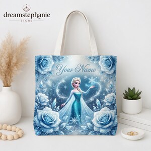 Op de afbeelding: Een witte draagtas met een blauwe illustratie van een prinses en de tekst "Your Name". Het ontwerp bevat blauwe rozen en sneeuwvlokken. De tas heeft cr&egrave;mekleurige handvatten en is geschikt voor het dragen van spullen.