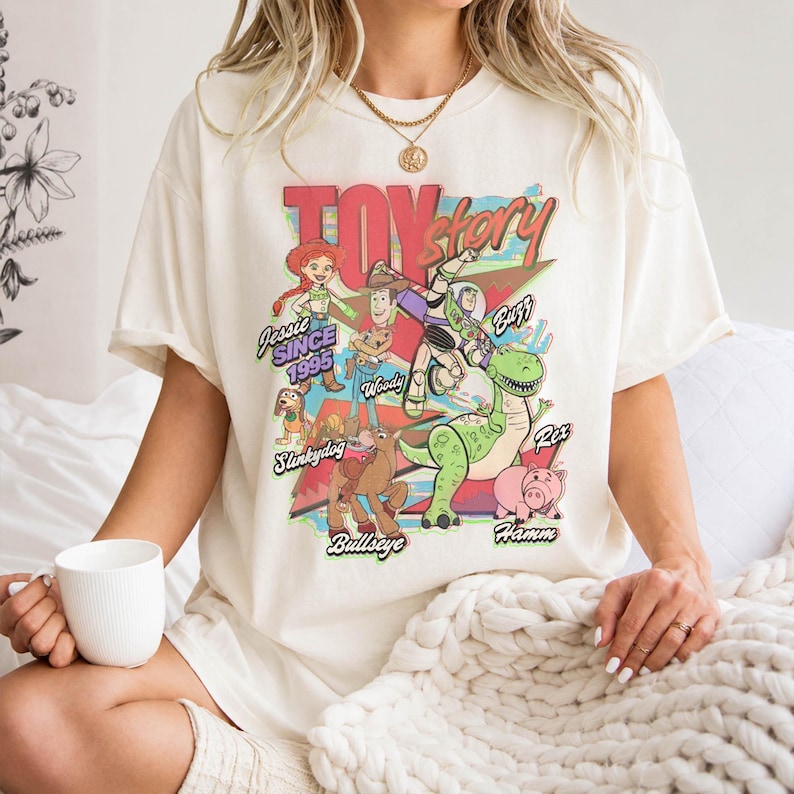 May include: An off-white short-sleeve t-shirt featuring a colorful graphic print of Toy Story characters including Jessie, Woody, Buzz Lightyear, Slinky Dog, Bullseye, Rex, and Hamm, with the text Toy Story and individual character names.