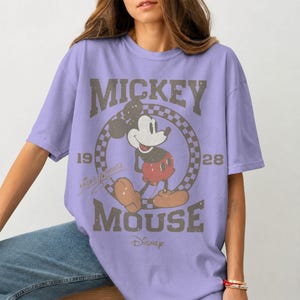 May include: A lilac-colored t-shirt featuring a distressed graphic of Mickey Mouse. The design includes the words "Mickey Mouse" and "Disney", along with the years "19" and "28". The shirt has a classic, vintage aesthetic.