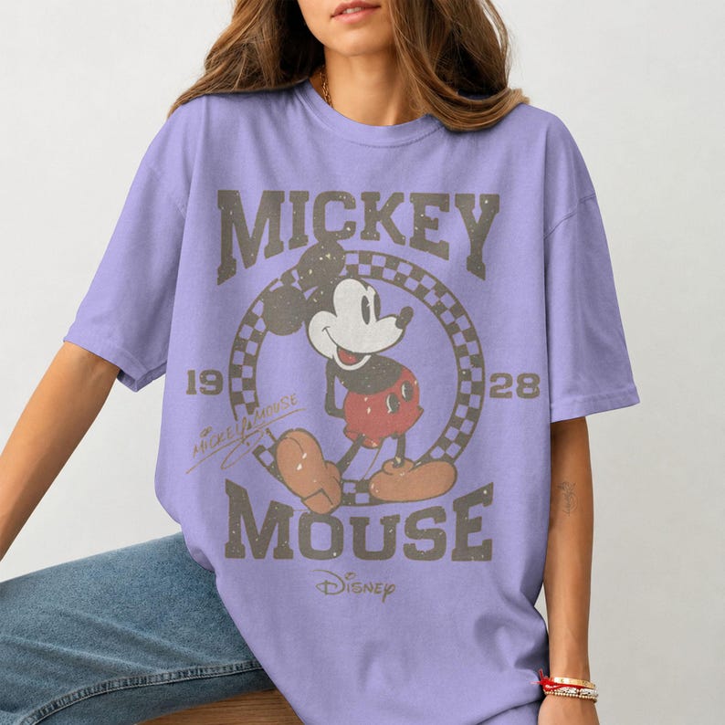 May include: A lilac-colored t-shirt featuring a distressed graphic of Mickey Mouse. The design includes the words "Mickey Mouse" and "Disney", along with the years "19" and "28". The shirt has a classic, vintage aesthetic.