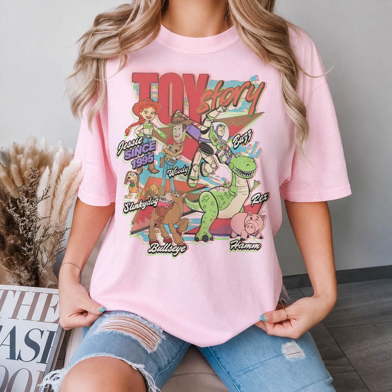 May include: A light pink t-shirt featuring a colorful graphic print of Toy Story characters including Woody, Buzz Lightyear, Jessie, Rex, Hamm, Slinky Dog, and Bullseye. The text reads Toy Story, Since 1995, and the names of the characters.