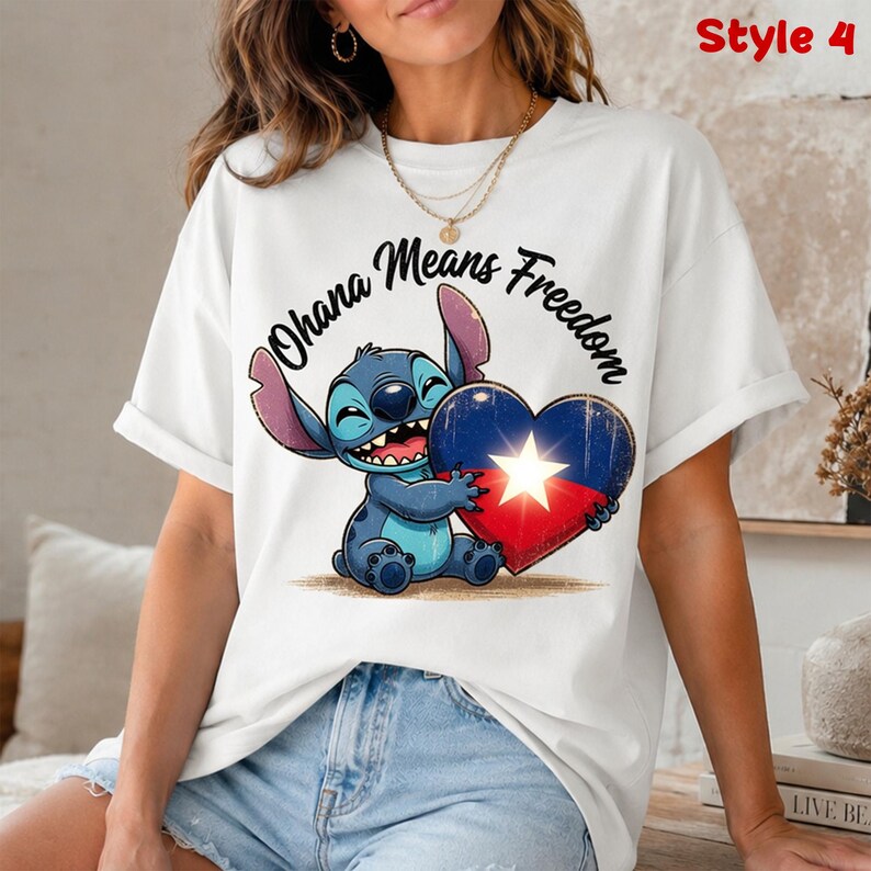 May include: White t-shirt featuring a cartoon character holding a heart-shaped design with a star. The text "Ohana Means Freedom" is above the character. The design is in shades of blue, red, and white.