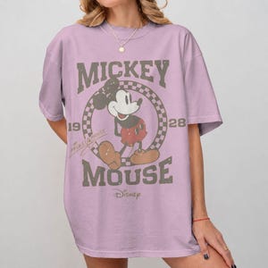 May include: A light purple t-shirt featuring a distressed graphic of Mickey Mouse. The design includes the words "Mickey Mouse" and "Disney" along with the years "1928" and "19". The cartoon character is in the center of a checkered circle.