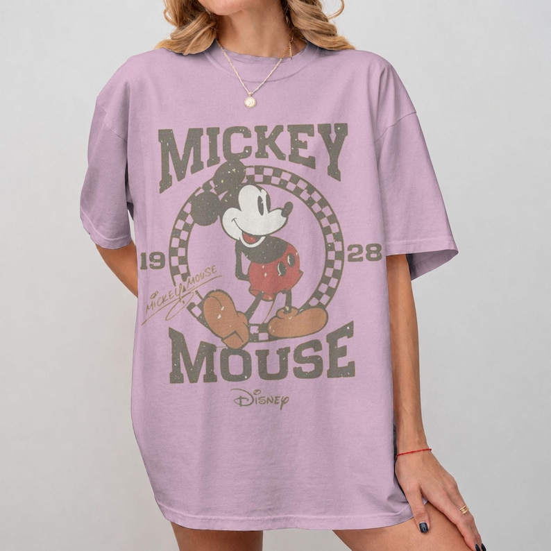 May include: A light purple t-shirt featuring a distressed graphic of Mickey Mouse. The design includes the words "Mickey Mouse" and "Disney" along with the years "1928" and "19". The cartoon character is in the center of a checkered circle.