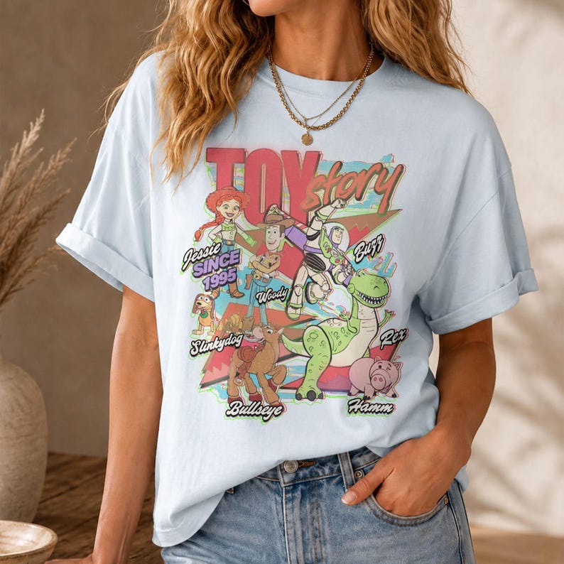 May include: A light blue short-sleeve t-shirt featuring a colorful graphic print of Toy Story characters including Jessie, Woody, Buzz Lightyear, Slinky Dog, Bullseye, Rex, and Hamm, with the text Toy Story and Jessie Since 1995 printed on the front.