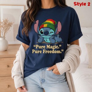 May include: Navy blue t-shirt featuring a cartoon character wearing a Rasta-style hat. The character is winking and resting its head on its hands. The shirt has the text "Pure Magic. Pure Freedom."
