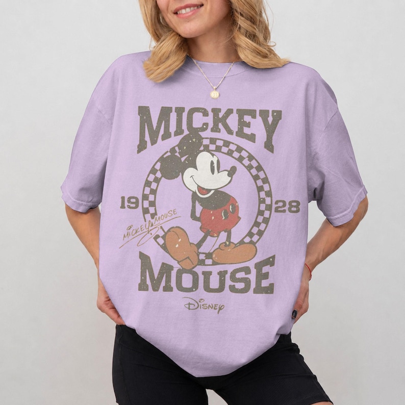 May include: A light purple t-shirt featuring a vintage Mickey Mouse graphic. The design includes the words "Mickey Mouse" and the year "1928" in a distressed, brown font. The Disney logo is at the bottom.
