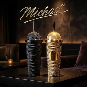 Op de afbeelding: Twee bekerglazen met studs, één zwart en één goudkleurig, met bijpassende deksels en rietjes, staan op een donkere ondergrond. Het woord "Michael" staat in gouden letters erboven. Elke beker heeft een cirkelvormig embleem met de naam "Michael".