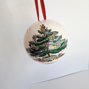May include: A spherical white ceramic Christmas bauble featuring a detailed illustration of a decorated evergreen tree with colourful ornaments, a figure at the top, and wrapped gifts at the base, suspended by a thin red ribbon loop.