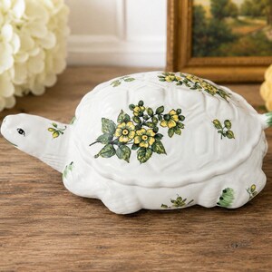 May include: A white ceramic turtle-shaped lidded box featuring a hand-painted floral pattern with yellow blossoms and green leaves across the shell and head. The turtle has a smooth, glossy finish and a sculpted, realistic form with a small tail.