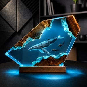May include: A decorative lamp featuring a humpback whale and two scuba divers suspended in a clear blue resin block. The piece is framed by natural wood edges with a glowing light effect and sits on a rectangular wooden base.