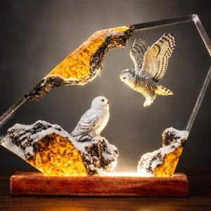 May include: A decorative lamp featuring two white snowy owls with black spots. One owl is perched on a snow-covered amber-colored rock, while the other is in flight. The scene is encased in a clear geometric resin frame on a rectangular wooden base.