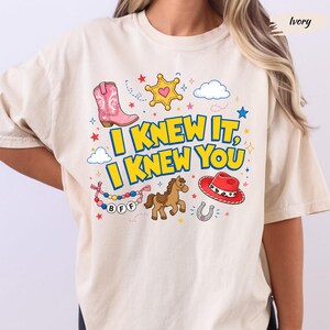 May include: An ivory short-sleeve t-shirt featuring a graphic print with the text I KNEW IT, I KNEW YOU in yellow letters. The design includes a pink cowboy boot, a yellow sheriff star, a brown horse, a red cowboy hat, and a beaded BFF bracelet.