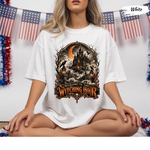 May include: A white t-shirt featuring a graphic print of a gothic castle under an orange crescent moon, surrounded by bare trees and ravens. The text "WELCOME TO THE WITCHING HOUR" is printed in orange and black at the bottom of the ornate frame.