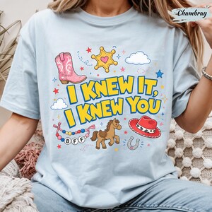 May include: A light blue t-shirt featuring the text I KNEW IT, I KNEW YOU in yellow with blue outlines. The graphic includes a pink cowboy boot, a yellow sheriff star, a brown horse, a red cowboy hat, a horseshoe, and a beaded BFF bracelet.