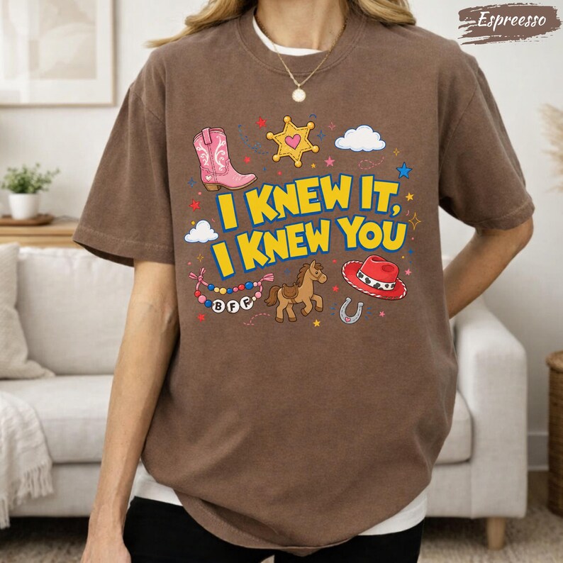 May include: A brown t-shirt featuring a graphic print with the text I KNEW IT, I KNEW YOU in yellow. The design includes a pink cowboy boot, a yellow star, a brown horse, a red cowboy hat, a horseshoe, and a beaded necklace with BFF charms.