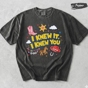 May include: A charcoal gray short-sleeve t-shirt featuring the yellow text I KNEW IT, I KNEW YOU. The graphic includes a pink cowboy boot, a yellow sheriff star, a brown horse, a red cowboy hat, a horseshoe, and a beaded necklace with BFF text.
